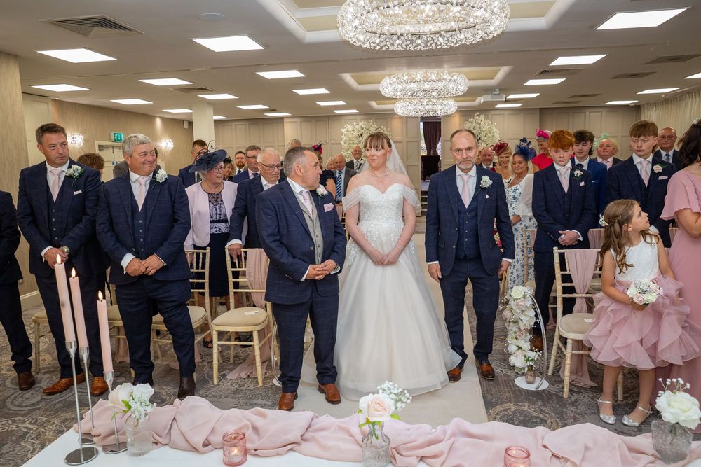 Bride walks down the aisle with escorts in an elegant chandelier-lit wedding ceremony venue.