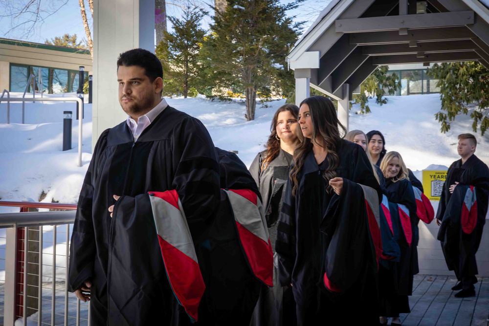 A group of graduates in black robes and stoles gather outside on a snowy winter day.