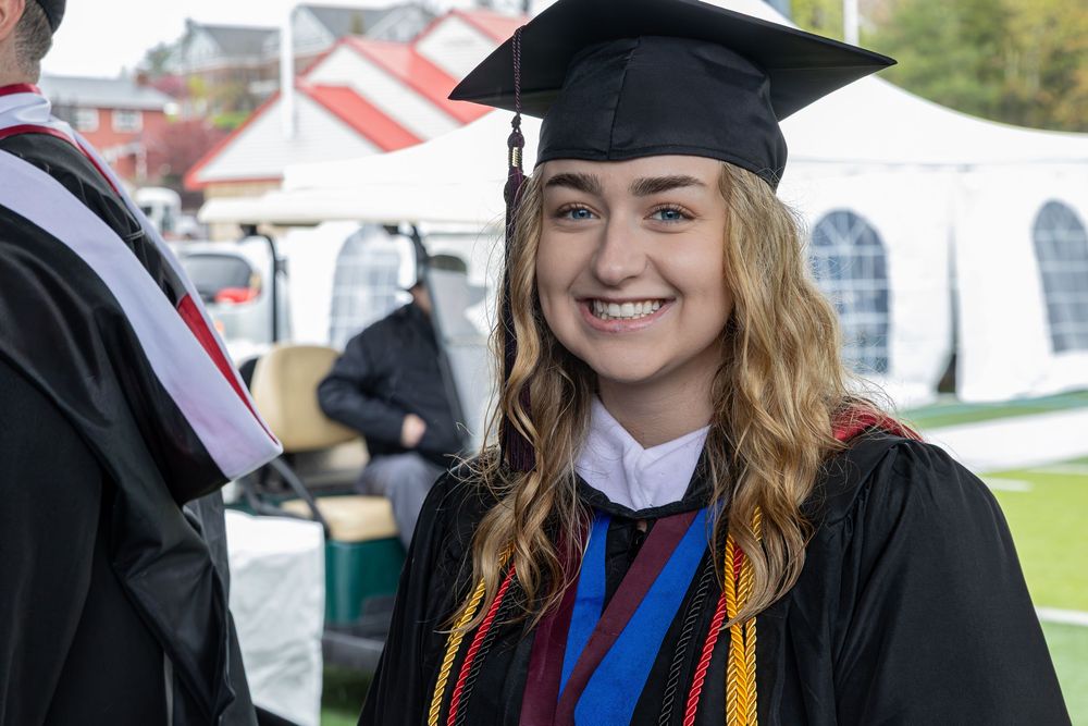 Graduate in traditional black cap and gown with honors cords at outdoor commencement ceremony.