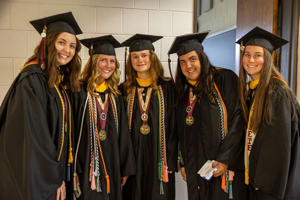 Five graduates in black caps and gowns with gold cords pose together at graduation.