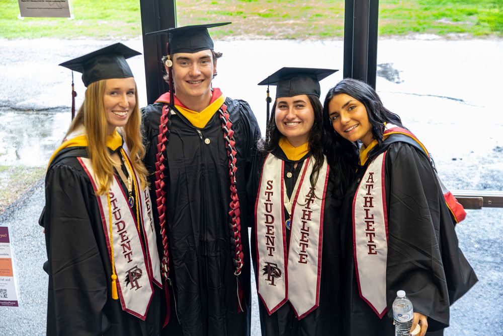 Four graduates in academic regalia and honor cords pose together outdoors.