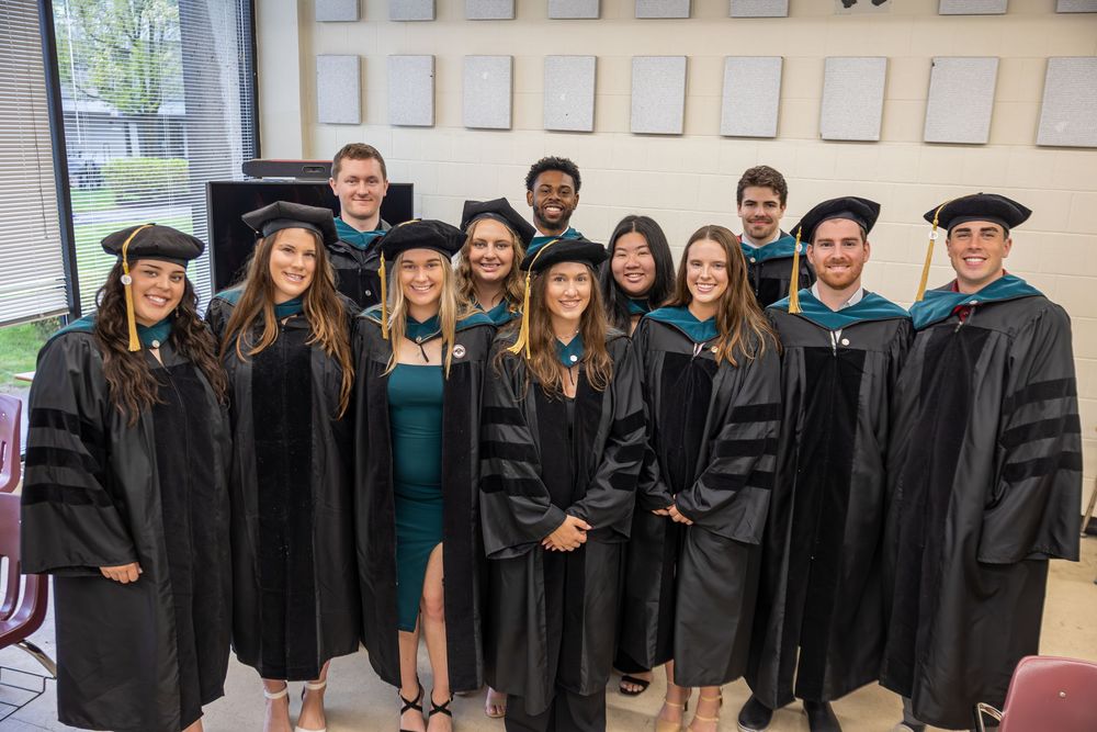 Group of graduates in academic regalia pose together in classroom setting.