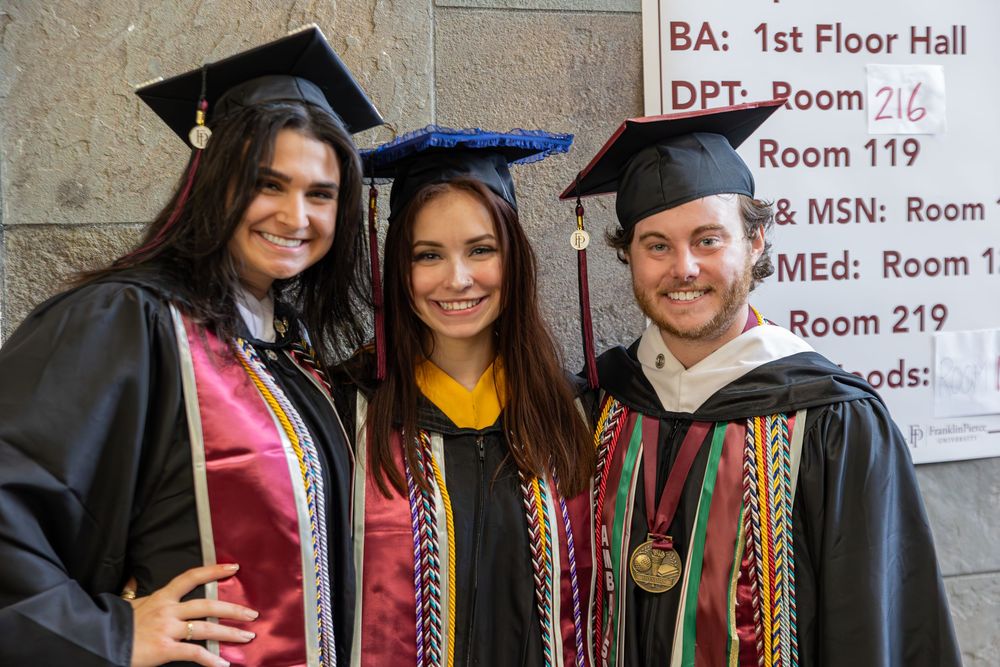 Three graduates in academic regalia pose in hallway near room directory sign.
