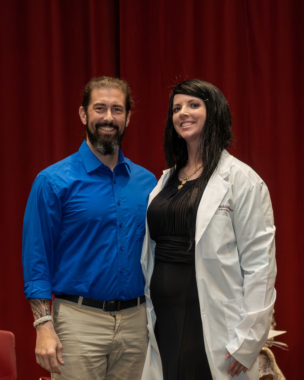 Two people pose in front of a red curtain backdrop, one wearing a blue shirt and khakis and another in professional attire.