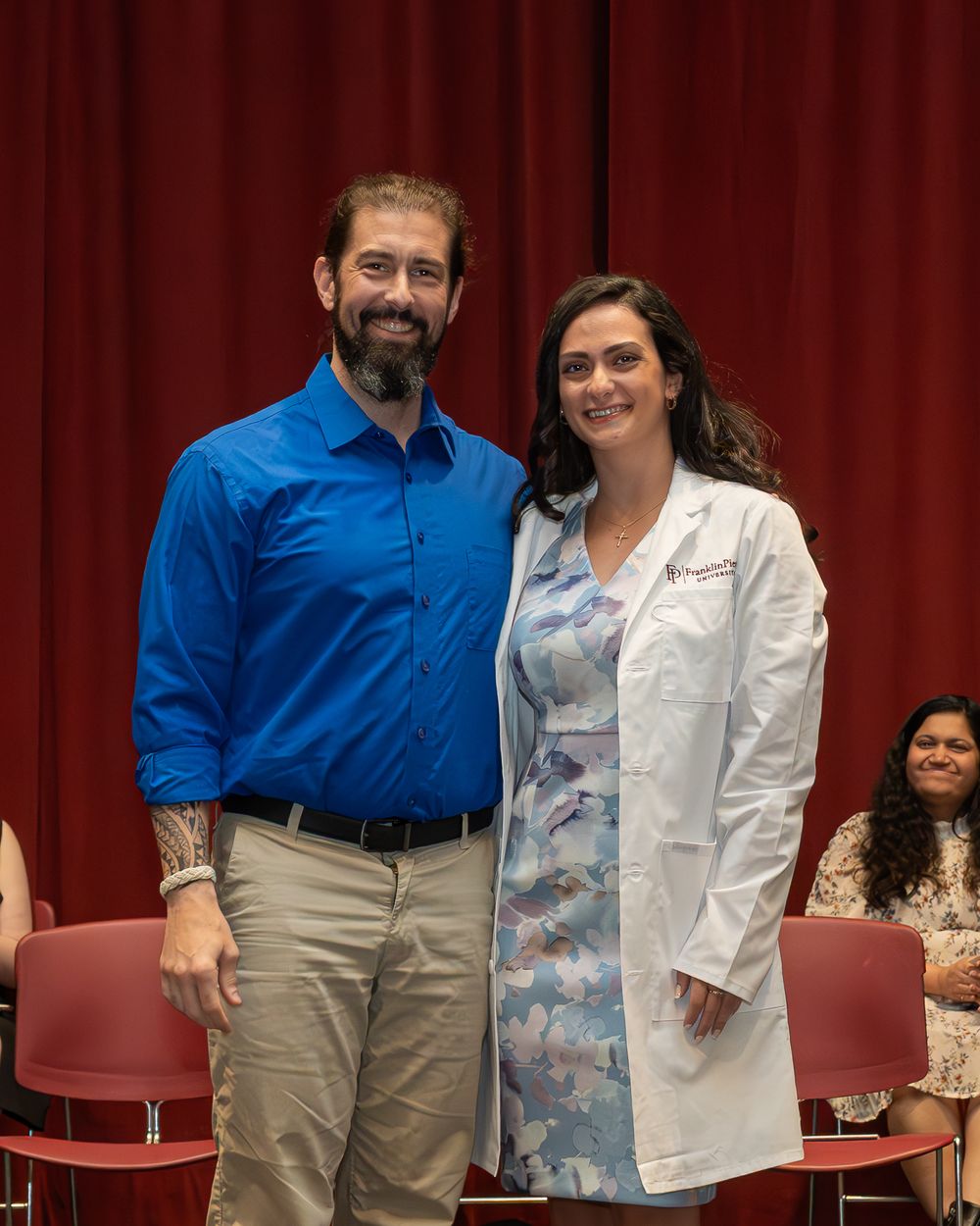Two people stand together against a red curtain, one in a blue shirt and another in a patterned white coat.