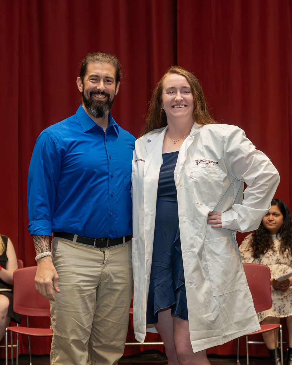 Professional in blue shirt poses with person in white medical coat against red curtain.