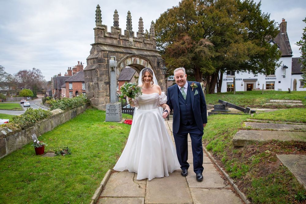Bride and father walk through church grounds with historic stone arch gateway behind them.