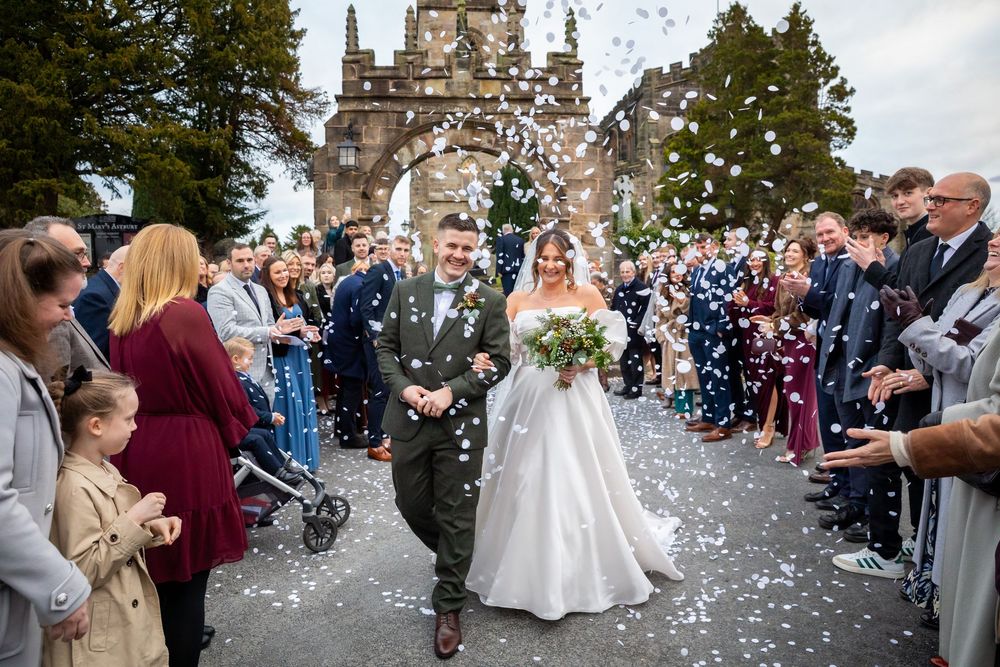 Bride and groom walk through confetti shower outside historic stone church arch with guests.