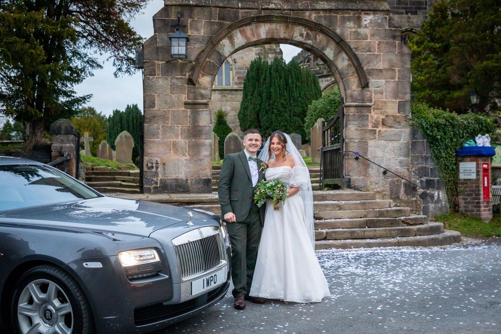 Bride and groom pose beside silver Rolls Royce outside historic stone church arch gateway.