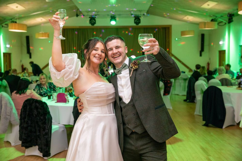 Bride and groom toasting with champagne glasses at their wedding reception in a green-lit ballroom.