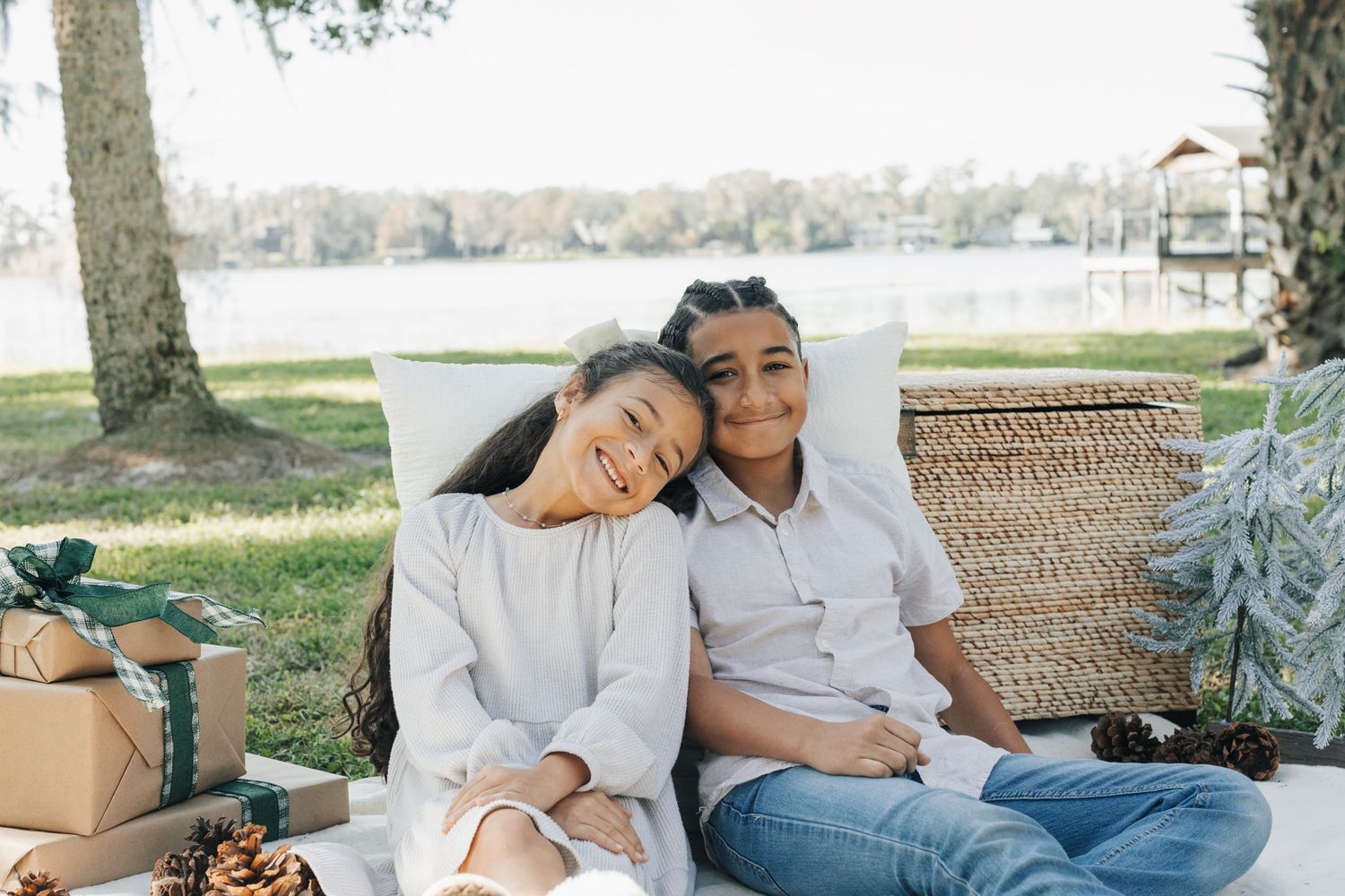 Two smiling children lean against each other on a picnic blanket beside a lake, next to wrapped gifts and pinecones.