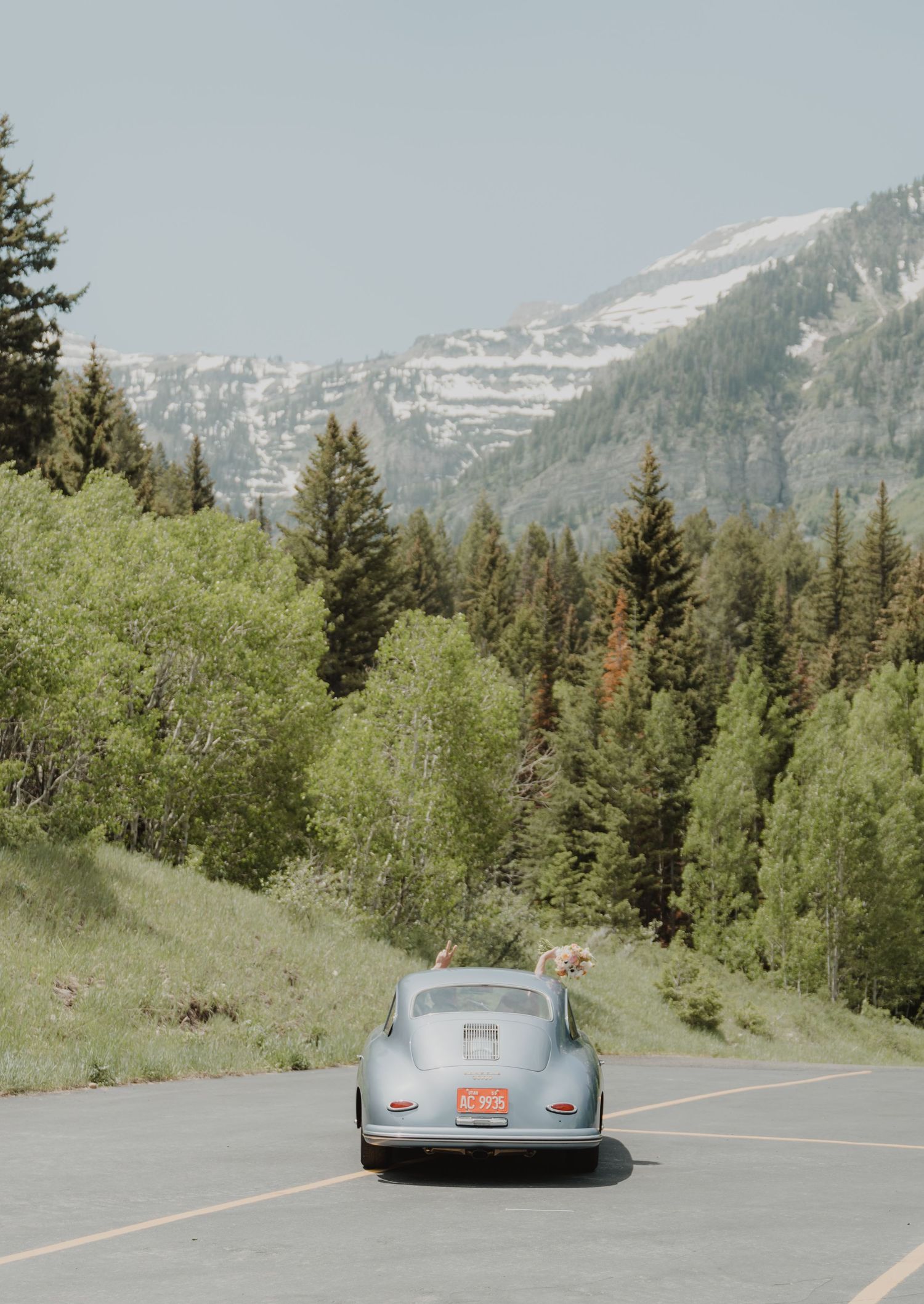 Classic silver car driving on a mountain road with snow-capped peaks and evergreen trees.