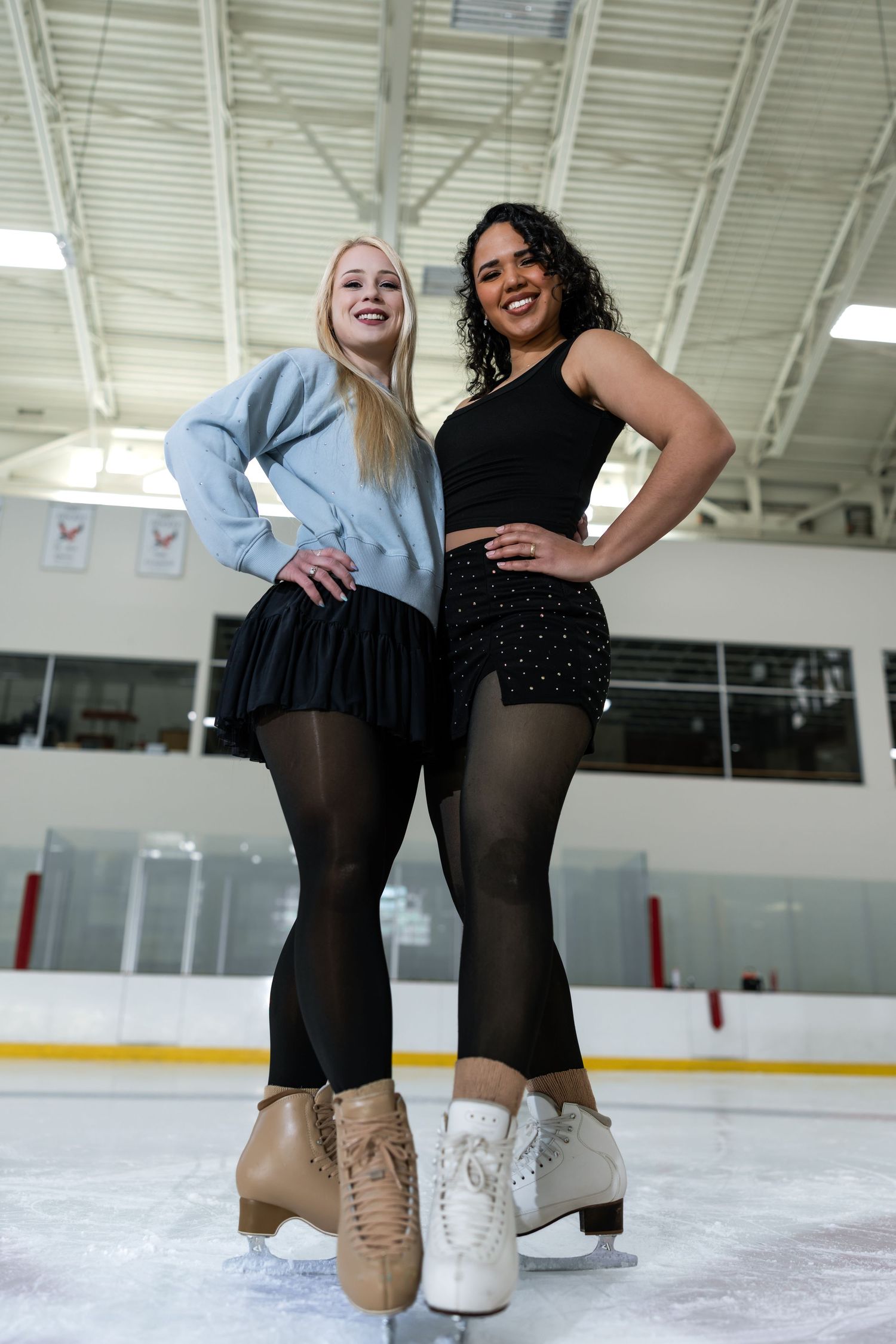 Two female figure skaters pose confidently on an indoor ice rink, wearing black skirts and tights with ice skates.