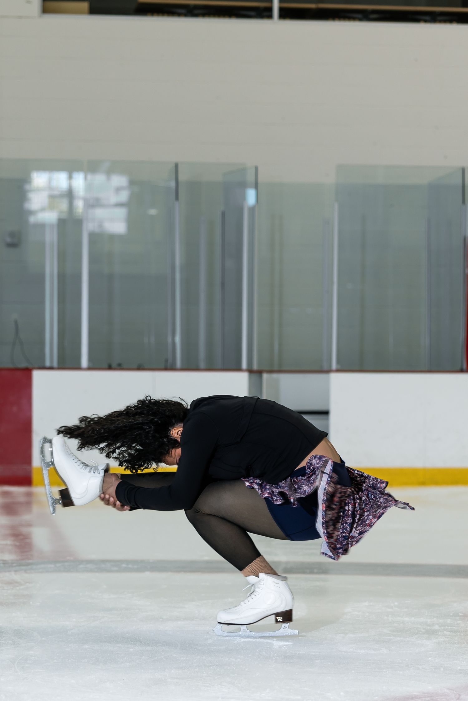 Female figure skater crouching low on ice rink performing a spin move wearing white skates and floral skirt.
