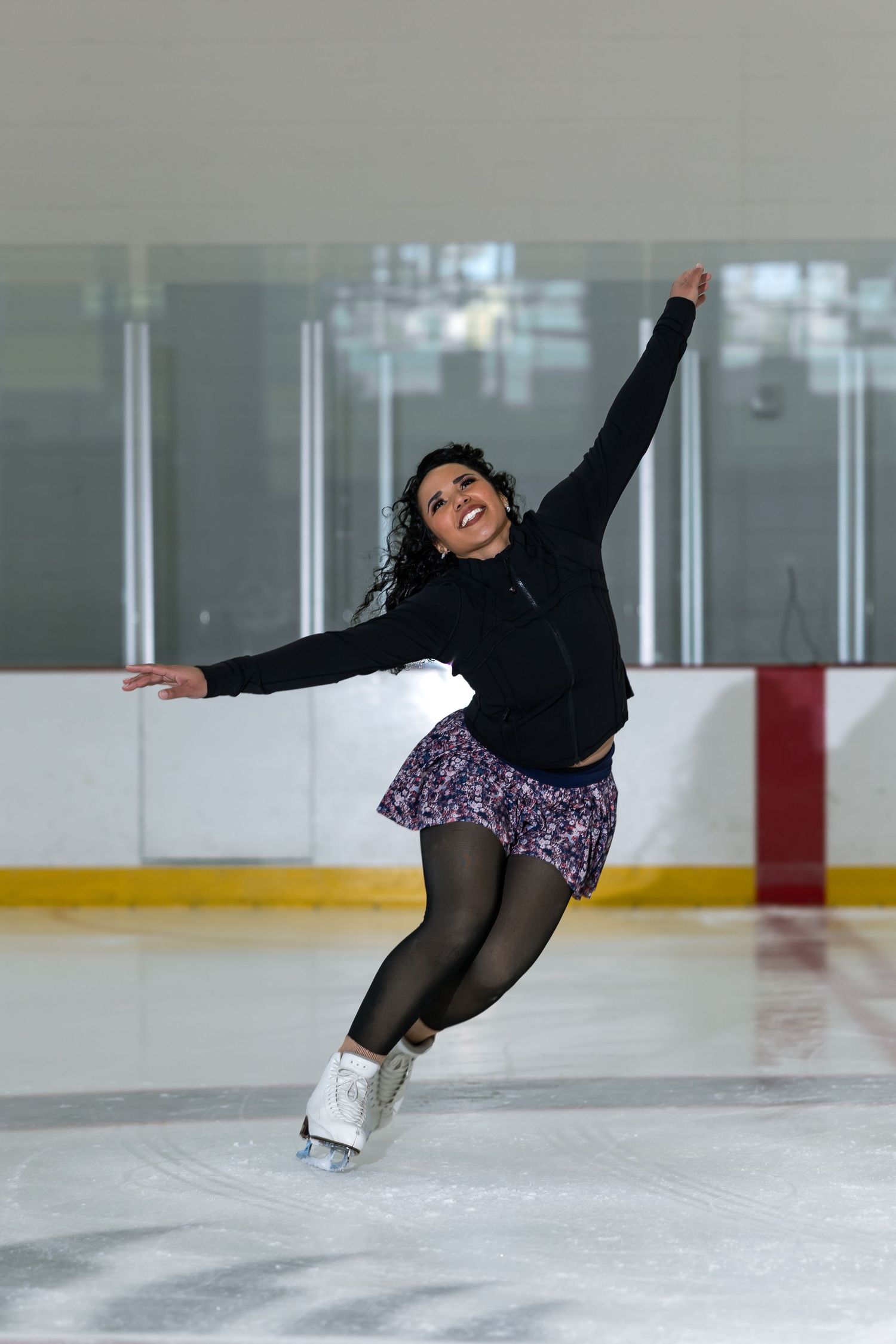 A female figure skater performs an elegant pose on an indoor ice rink, wearing a black jacket and floral skirt.