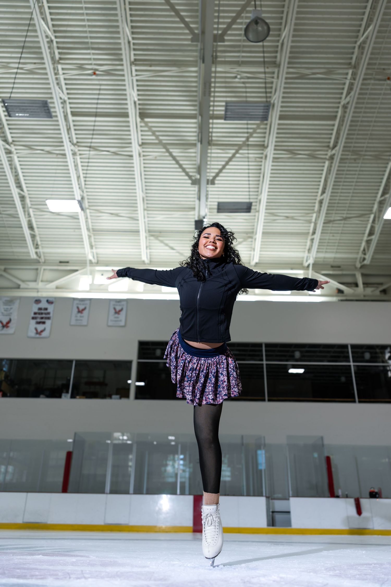 Young female figure skater jumping gracefully on indoor ice rink, wearing black jacket and purple sparkle skirt.