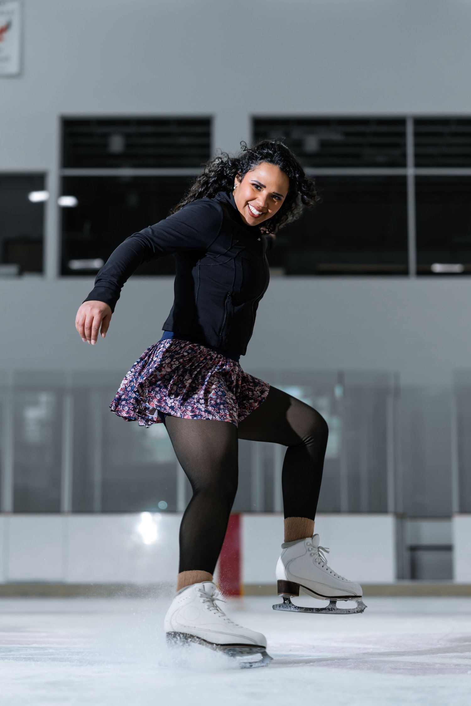 Female figure skater performing on indoor ice rink wearing black top and floral skirt.