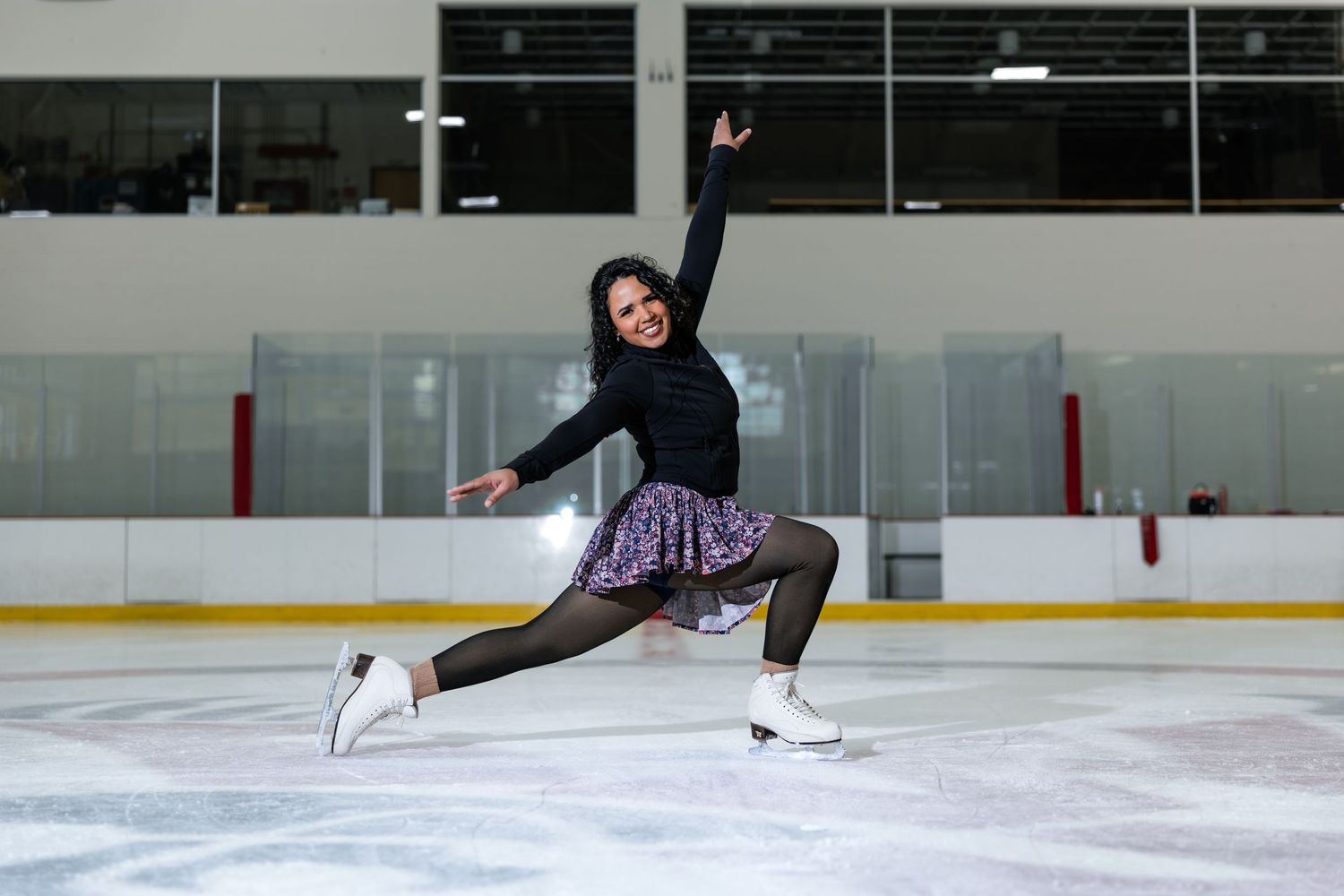Female figure skater performs a graceful split pose on ice rink wearing black top and purple skating skirt.