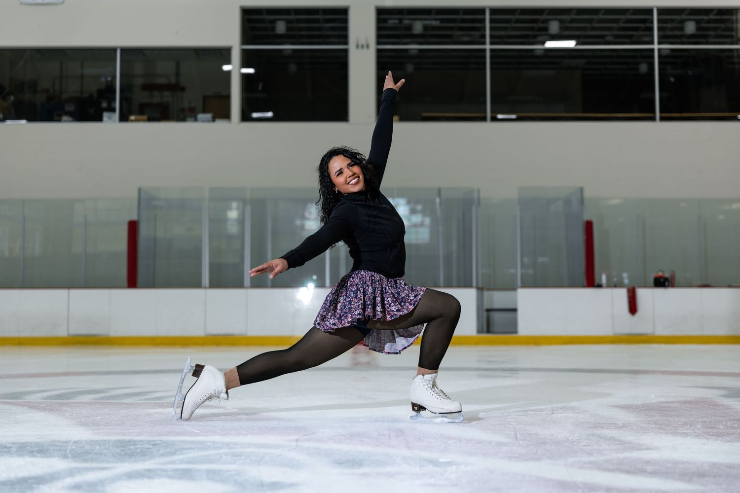 Female figure skater in black top and purple skirt performs graceful split jump pose on indoor ice rink.
