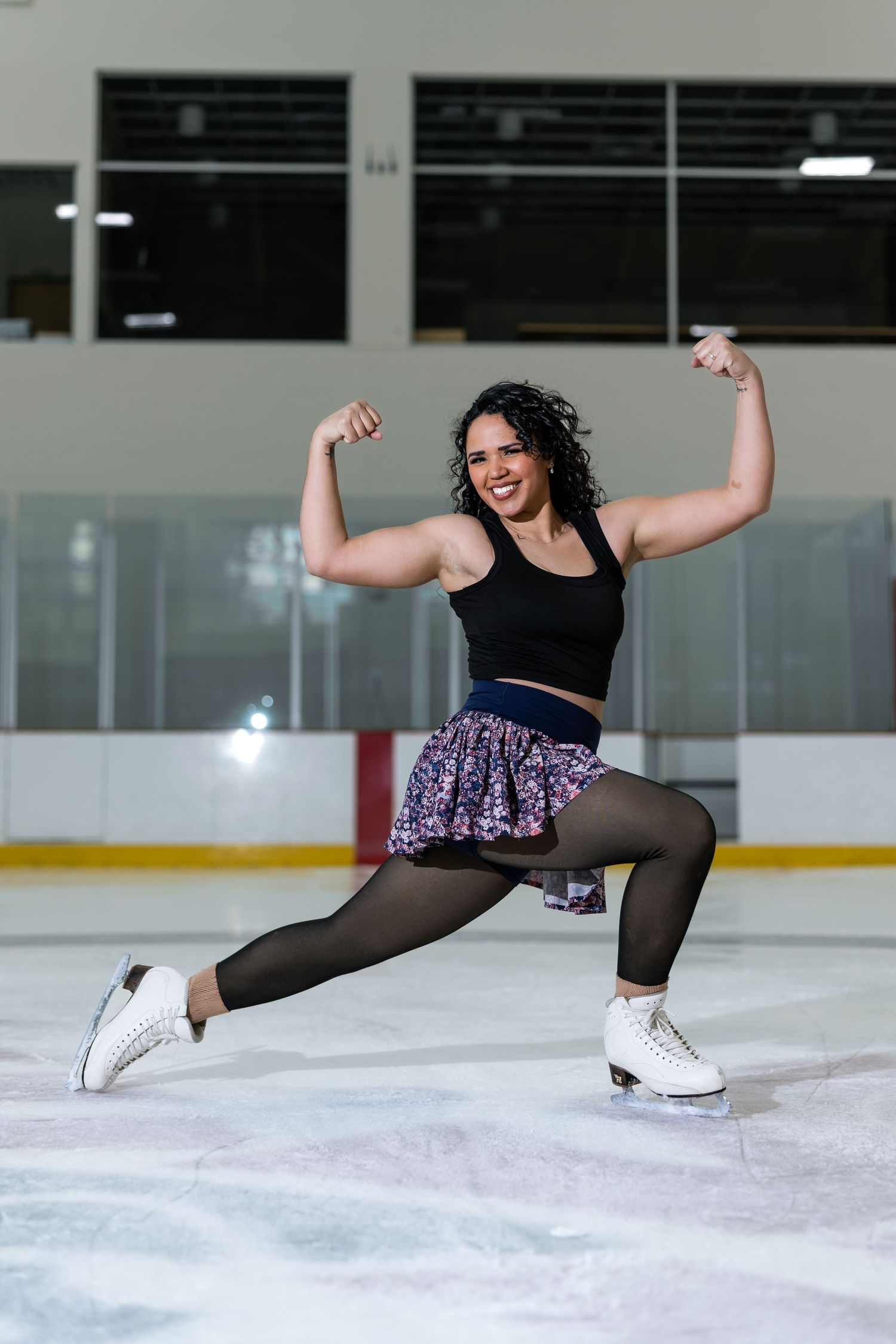 Confident female figure skater flexing muscles on ice rink wearing white skates and floral skirt.