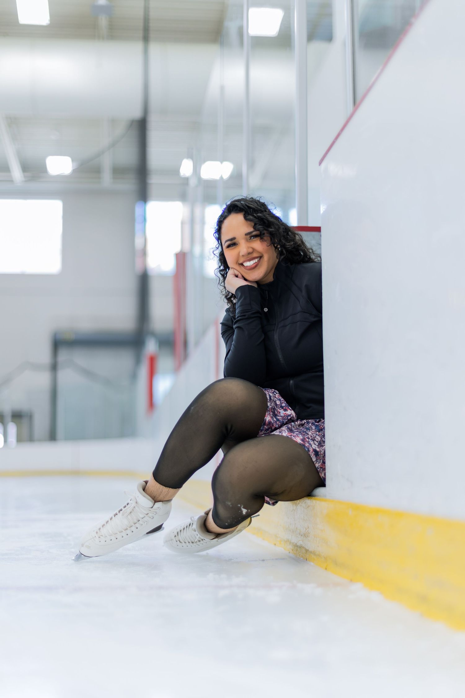 Woman in figure skates sitting on ice rink floor, smiling in black top and floral skirt.