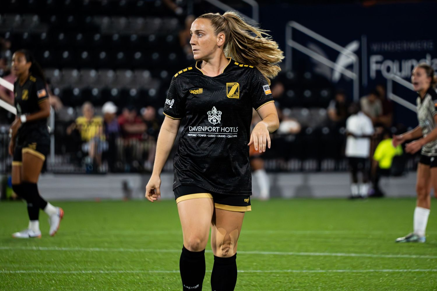 A soccer player in a black uniform kicks the ball during a nighttime match on an illuminated field.