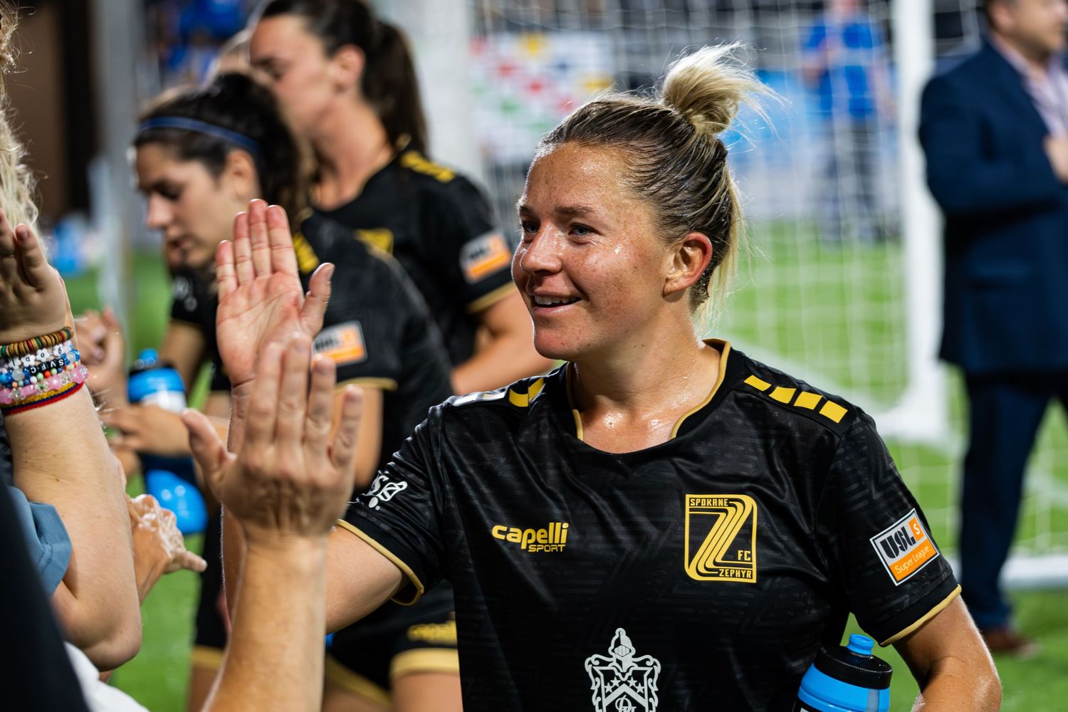 Players celebrate with fans after a soccer match, with black and yellow uniforms visible in the crowd.