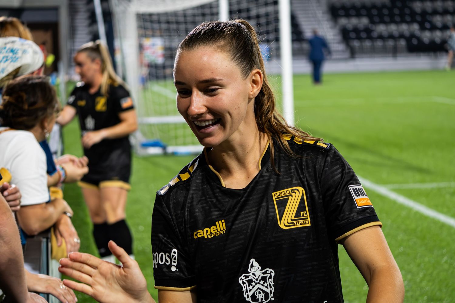 Soccer player in black and yellow uniform smiles while interacting with fans after a match.
