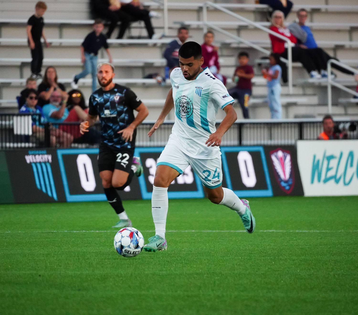 Soccer player in white uniform dribbles ball during match on professional field with stadium seating visible.