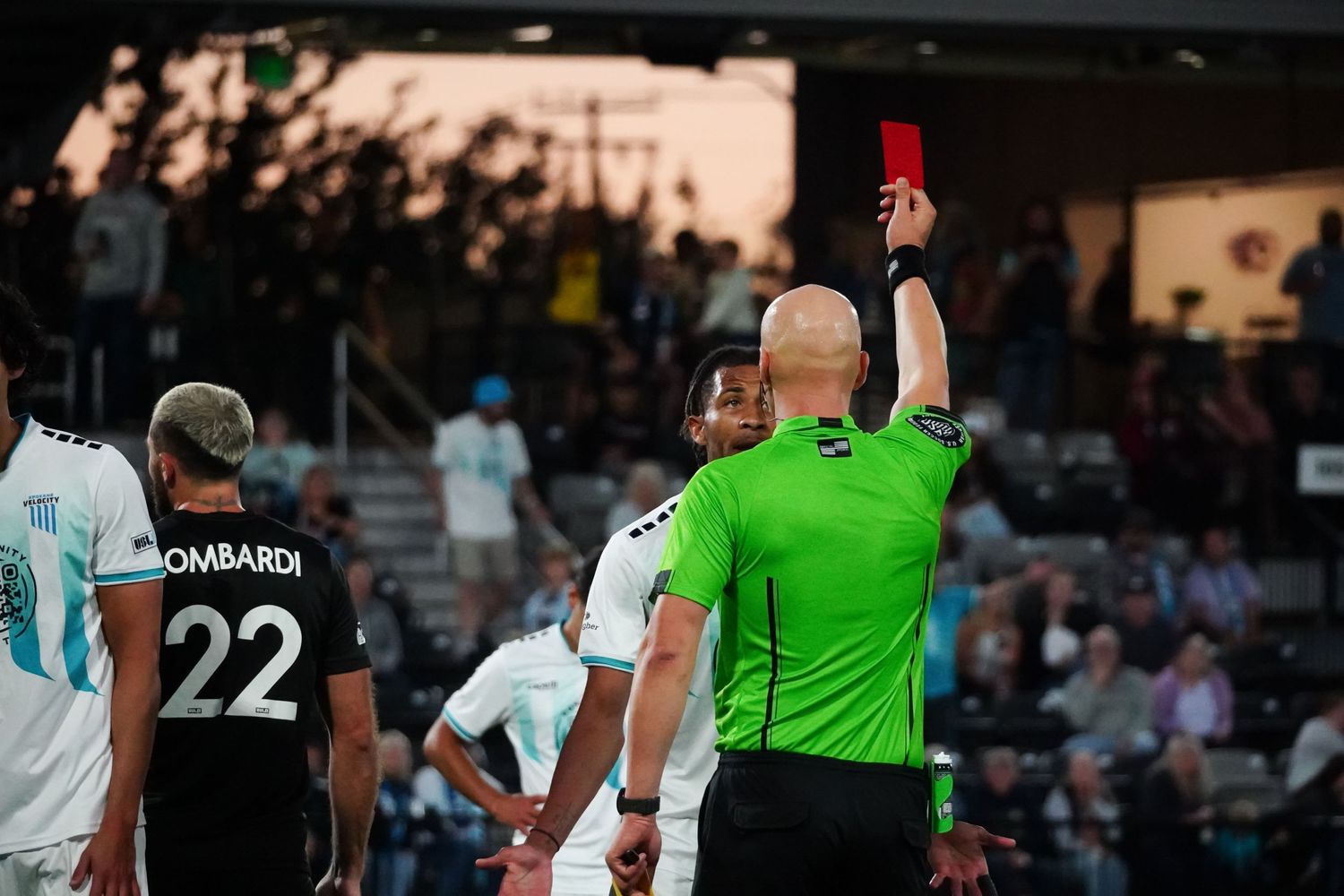 Soccer referee shows a red card to a player during a match while wearing a bright green uniform.