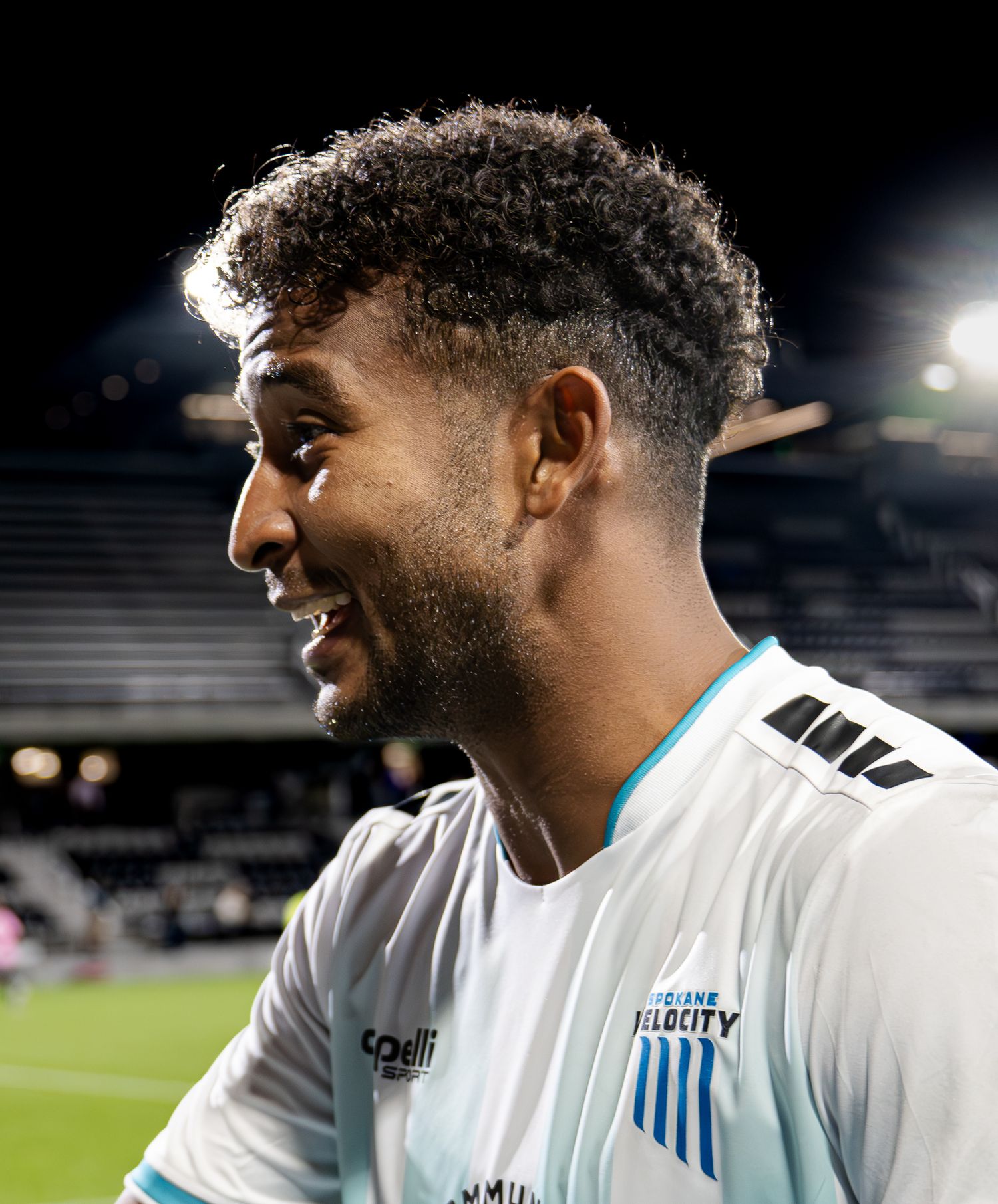 Professional soccer player in a white jersey smiles while on the field during a nighttime match.