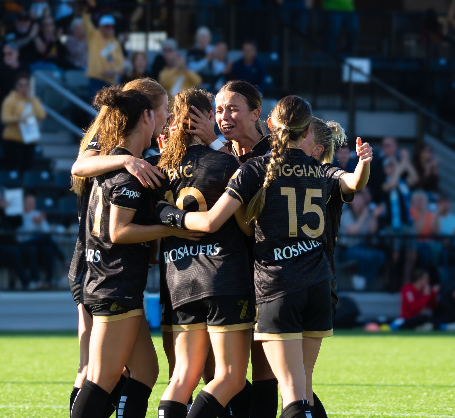 Female soccer players celebrate together in black uniforms on a sunny field during a match.