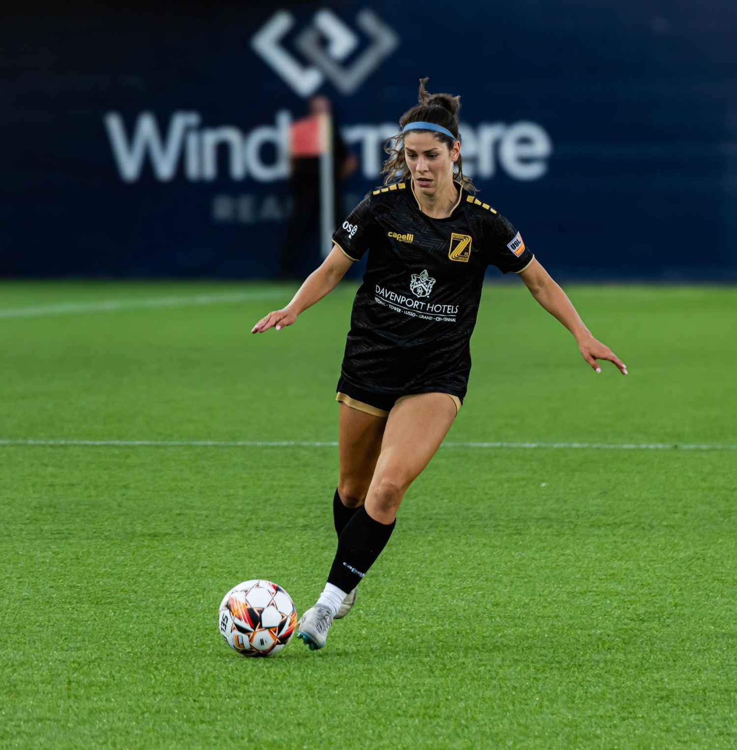 Soccer player in black uniform dribbles ball across green turf field during competitive match.