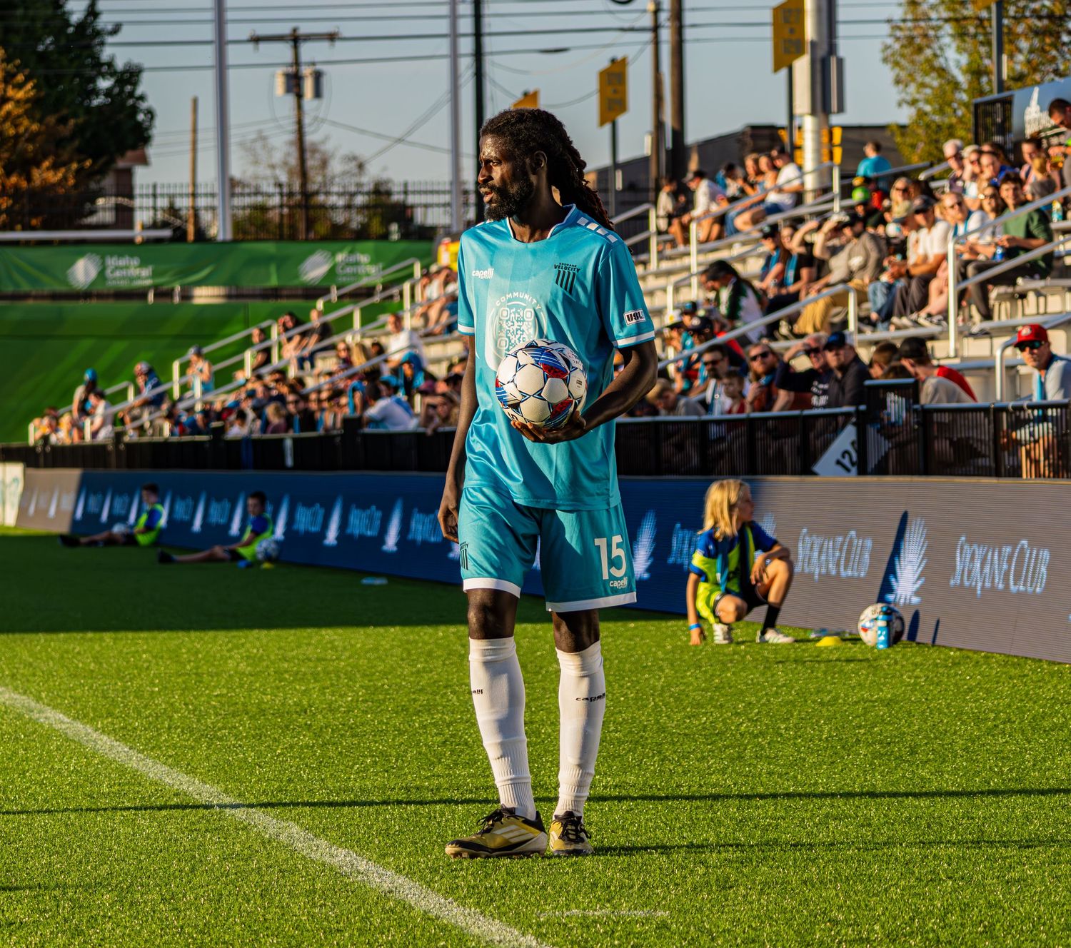 Soccer player in light blue jersey holds ball on sideline during match with crowd in background at outdoor stadium.