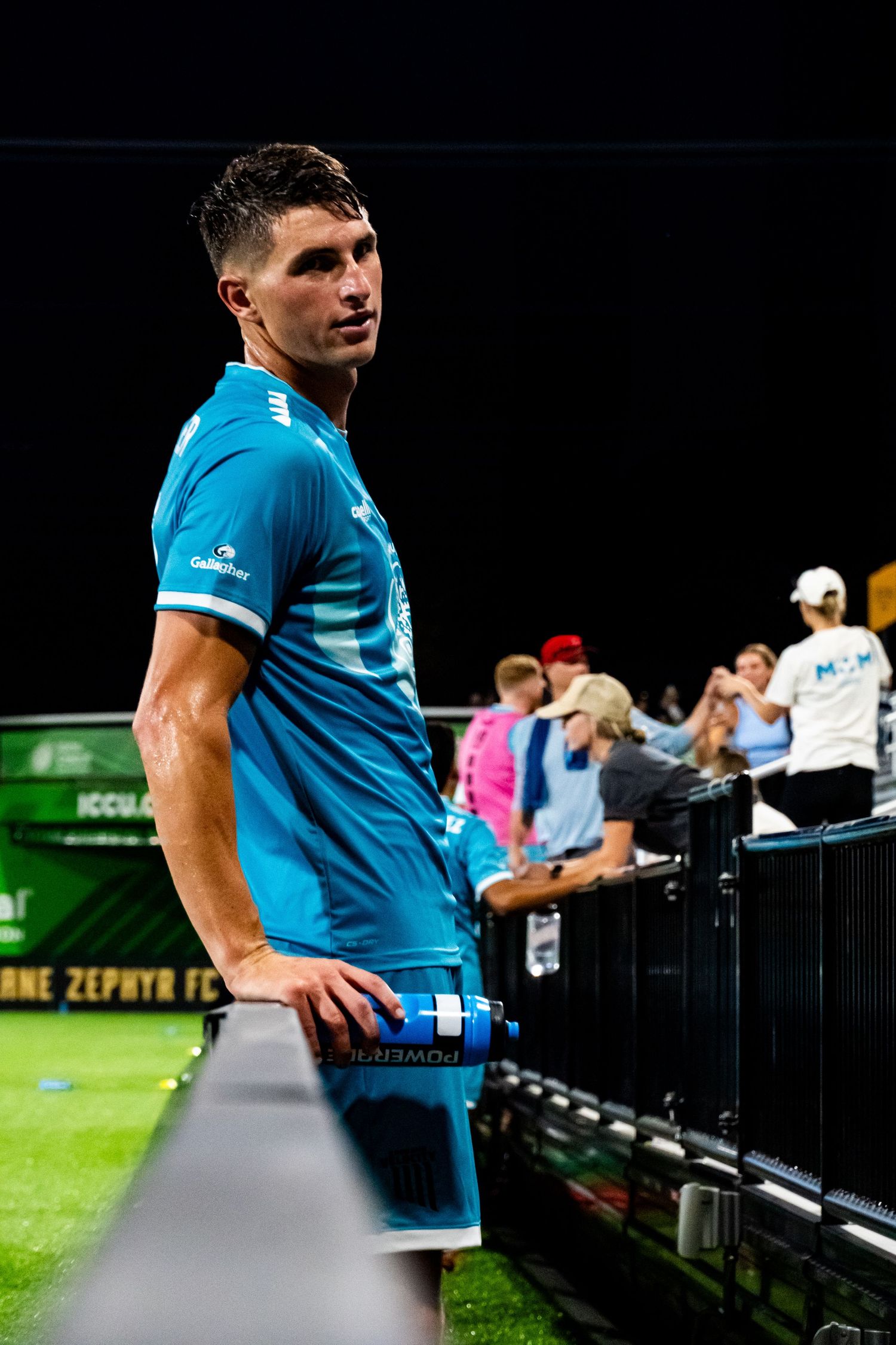 Soccer player in bright blue jersey stands near sideline barrier during nighttime match with spectators in background.