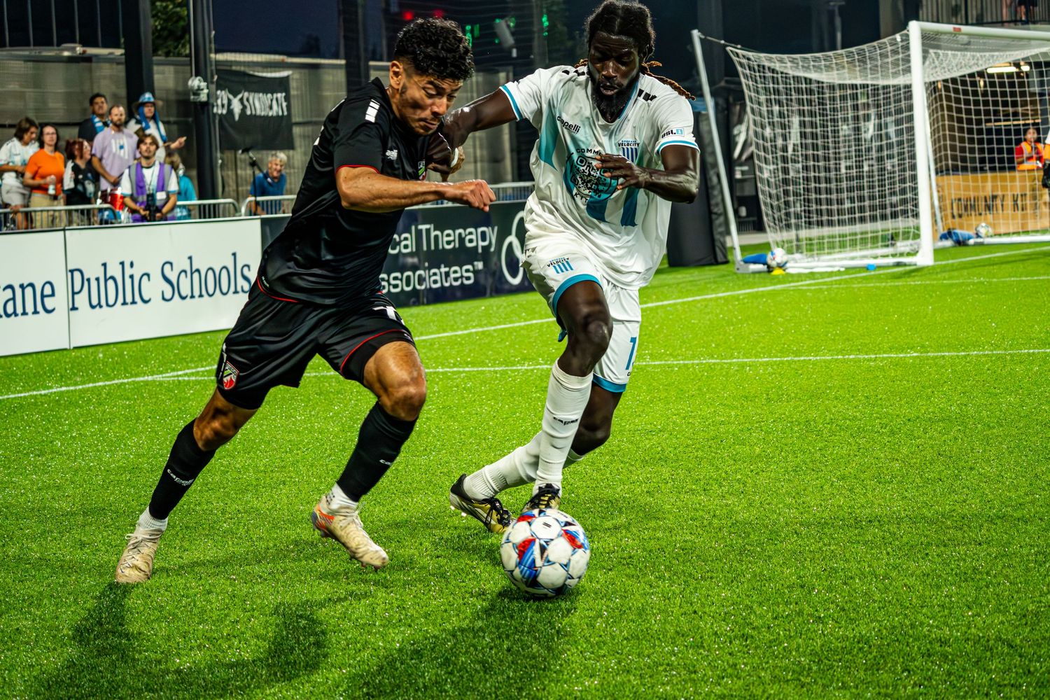 Two soccer players battle for possession of the ball during a nighttime match on an artificial turf field.