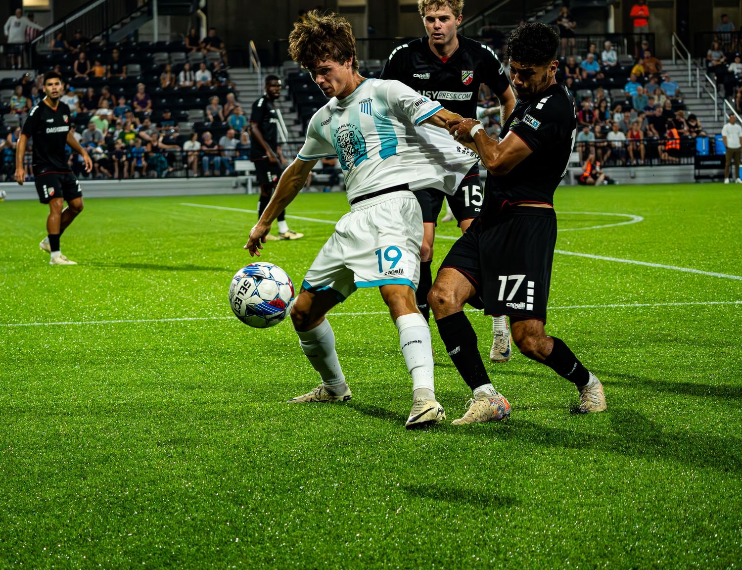 Soccer players from opposing teams battle for possession of the ball during an intense match on a floodlit field.