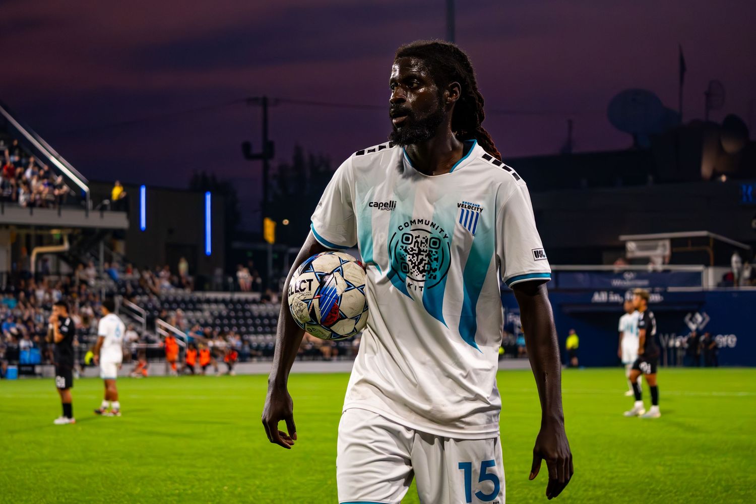 Soccer players in white jerseys compete during a nighttime match at a stadium under bright floodlights.