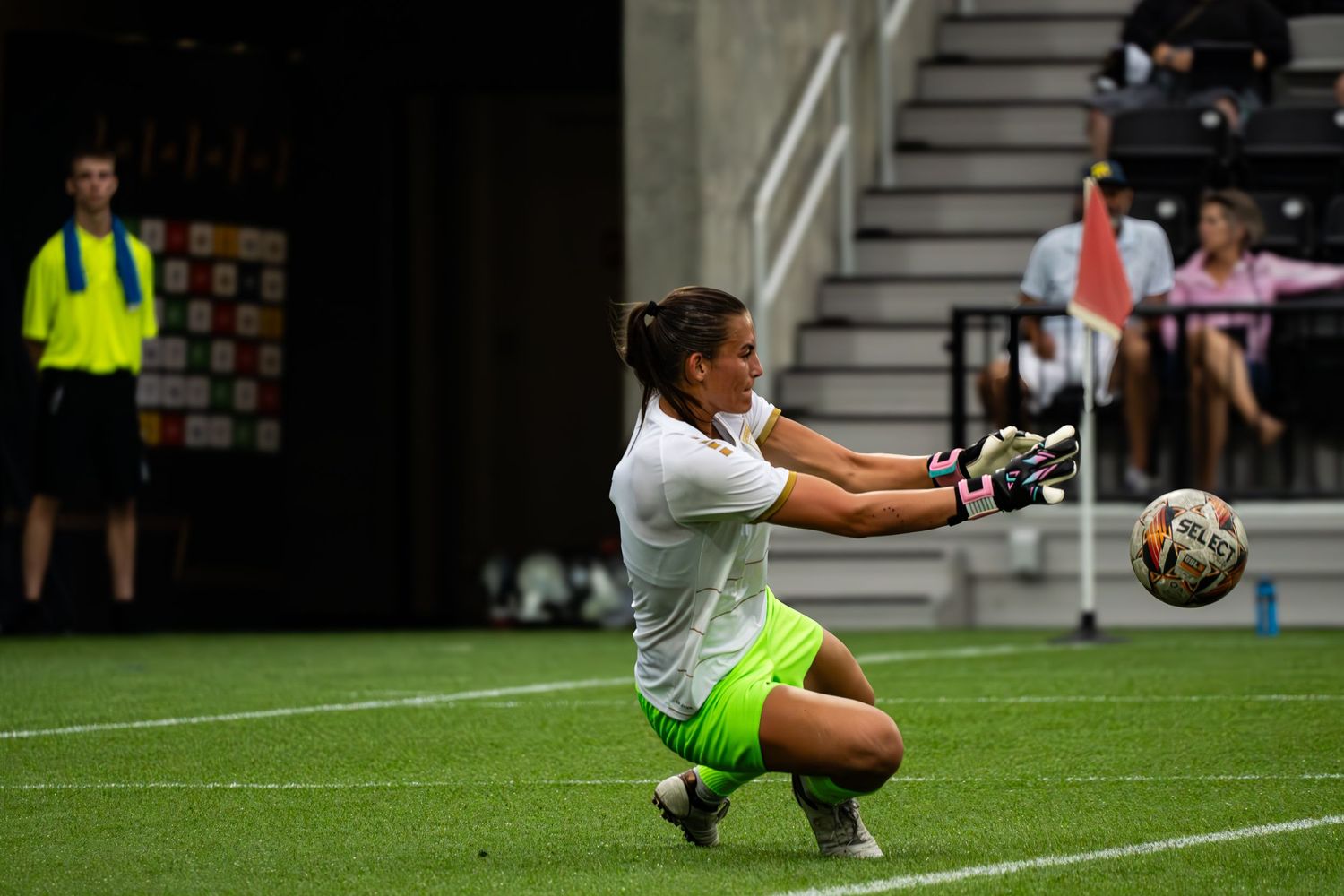 Soccer goalkeeper in white jersey and neon shorts makes diving save during indoor match.