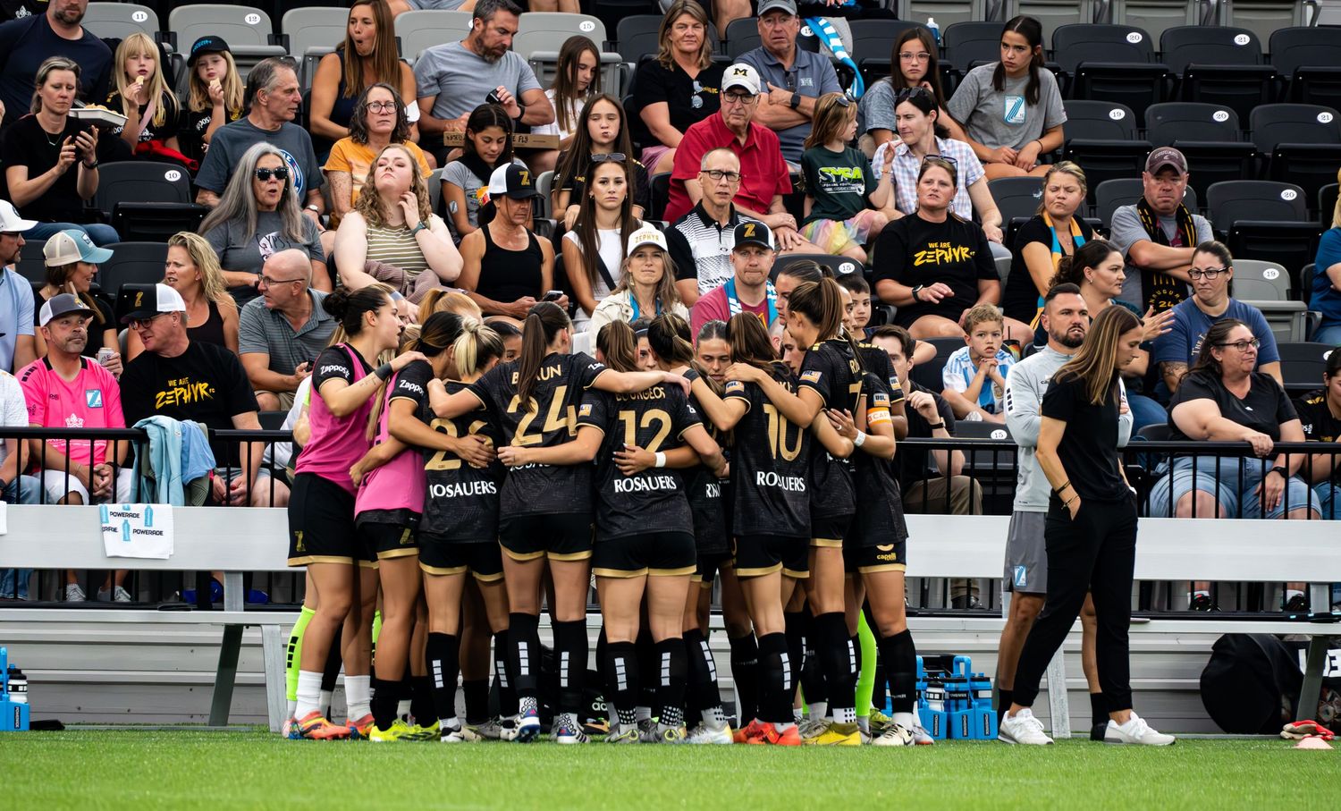 A sports team huddles together in black uniforms on a field while spectators watch from stadium seating in the background.