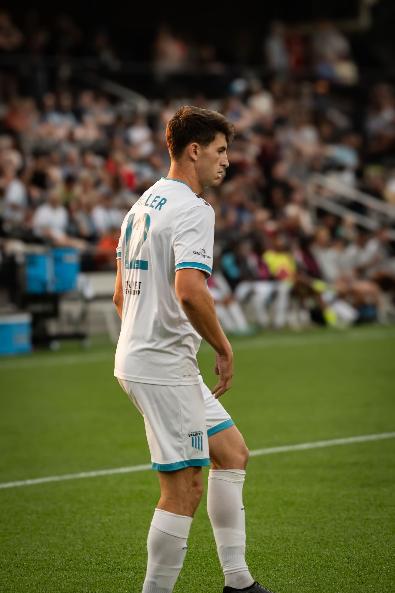 Soccer player in white uniform with turquoise accents stands on the sideline during a match.
