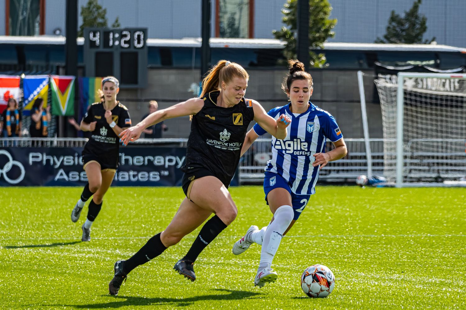 Two soccer players compete for the ball during a match on a sunny day at an outdoor stadium field.