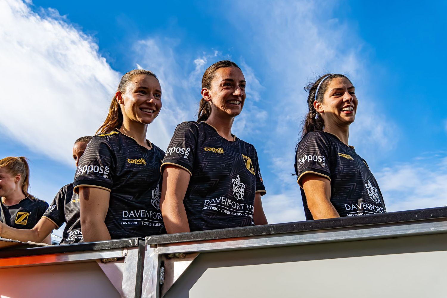 A group of athletes in matching black uniforms stand together against a bright blue sky background.