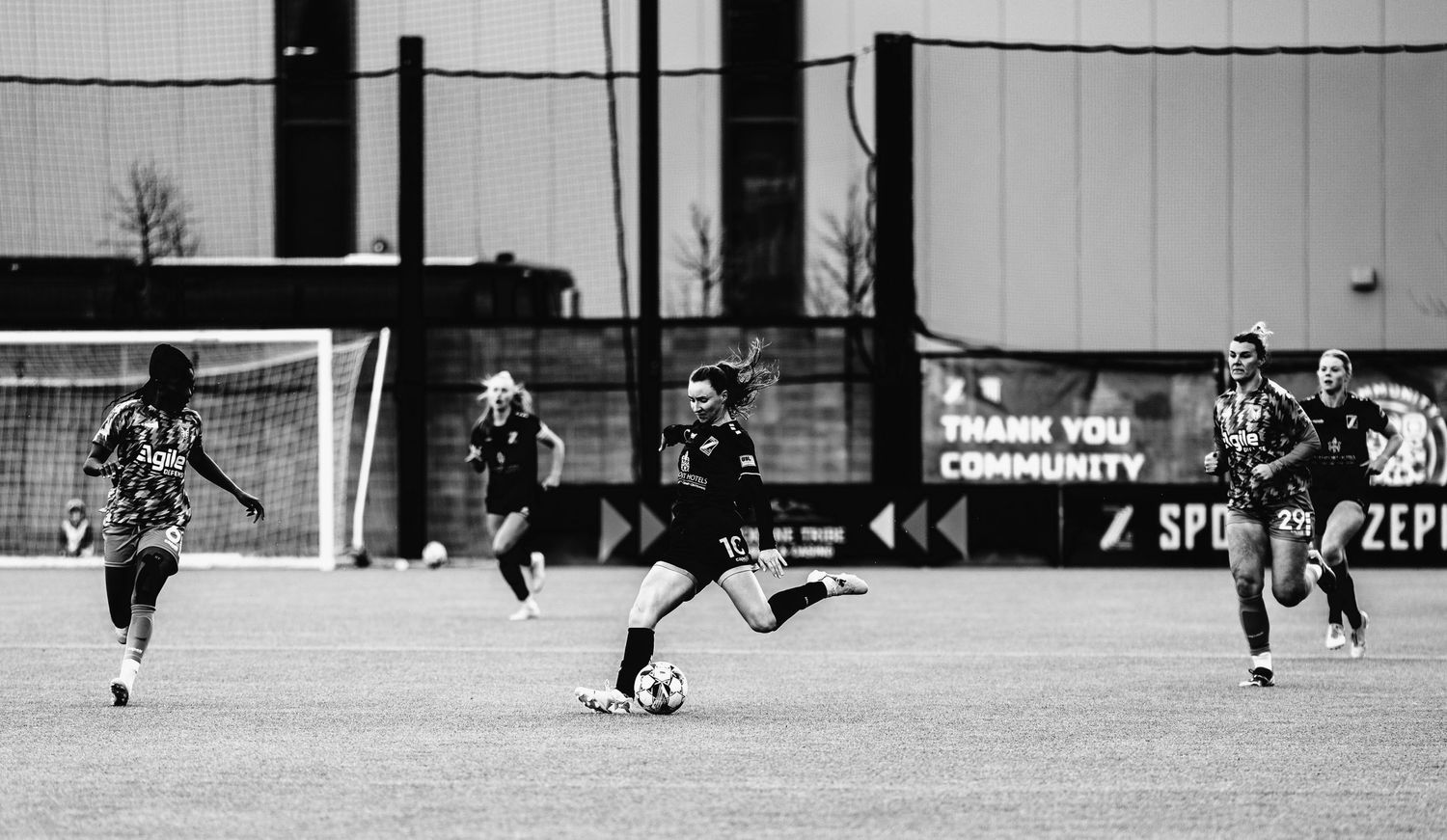 Black and white photo of soccer players in action on a field during a match, with a goal visible in the background.