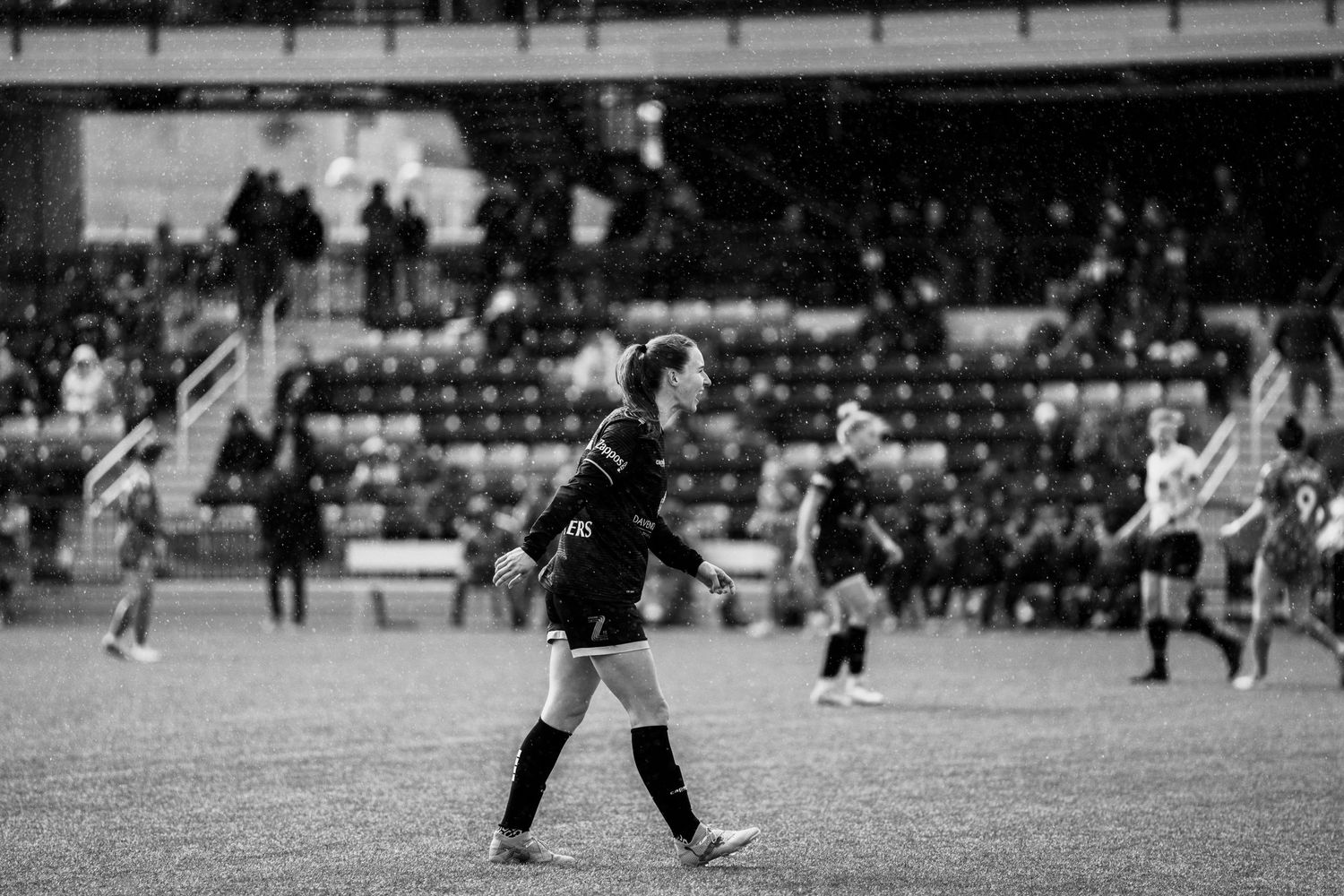 A soccer player in uniform stands on a field during a match with spectators visible in the background.