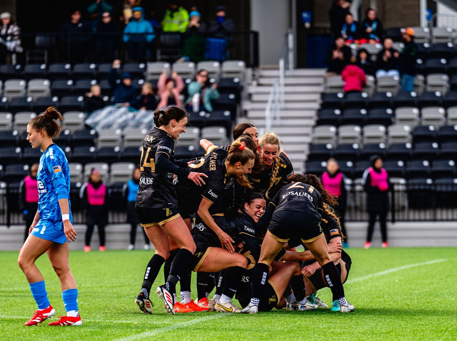 A soccer match in progress with players engaged in a scrum formation on a grass field surrounded by spectators.