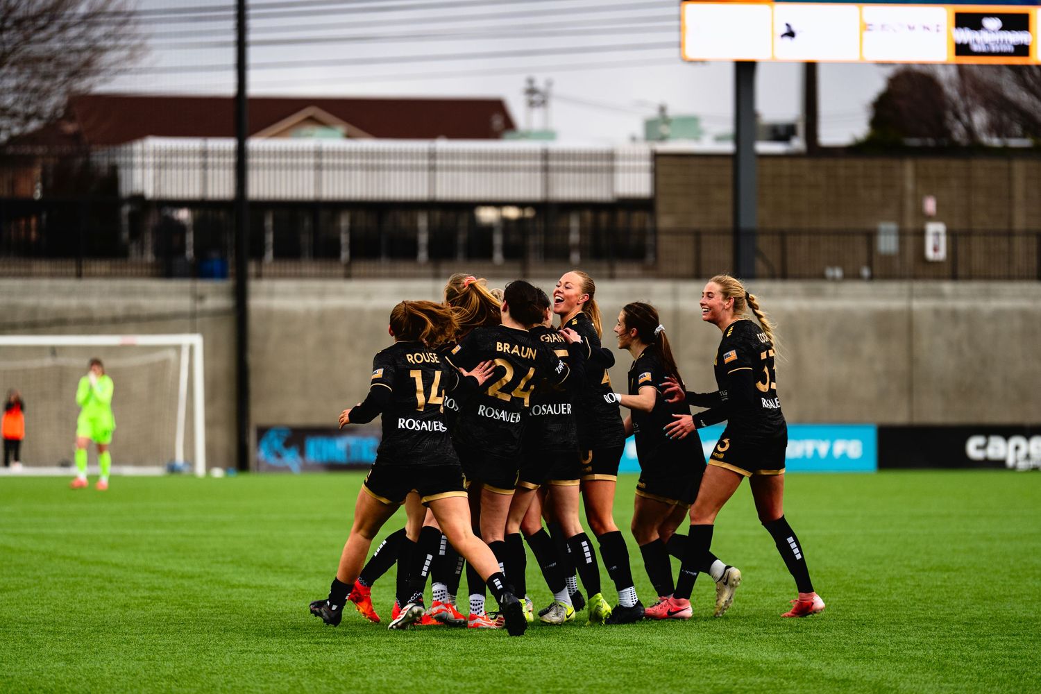Soccer team players huddle together in celebration on a grass field during a match at dusk.