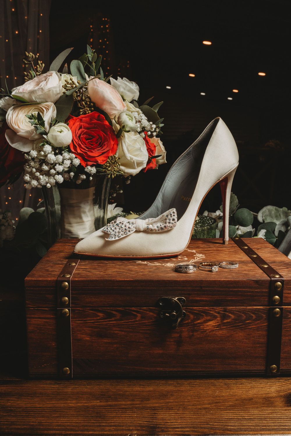 White bridal shoe with crystal embellishment next to floral arrangement on wooden chest.