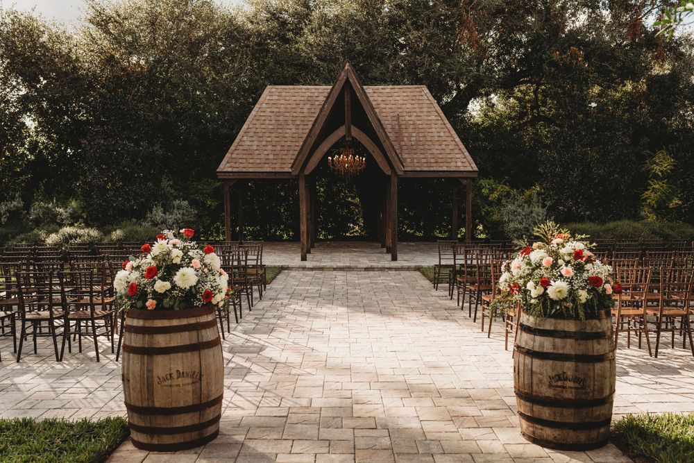 Rustic outdoor wedding ceremony setup with wooden gazebo and wine barrel flower arrangements.