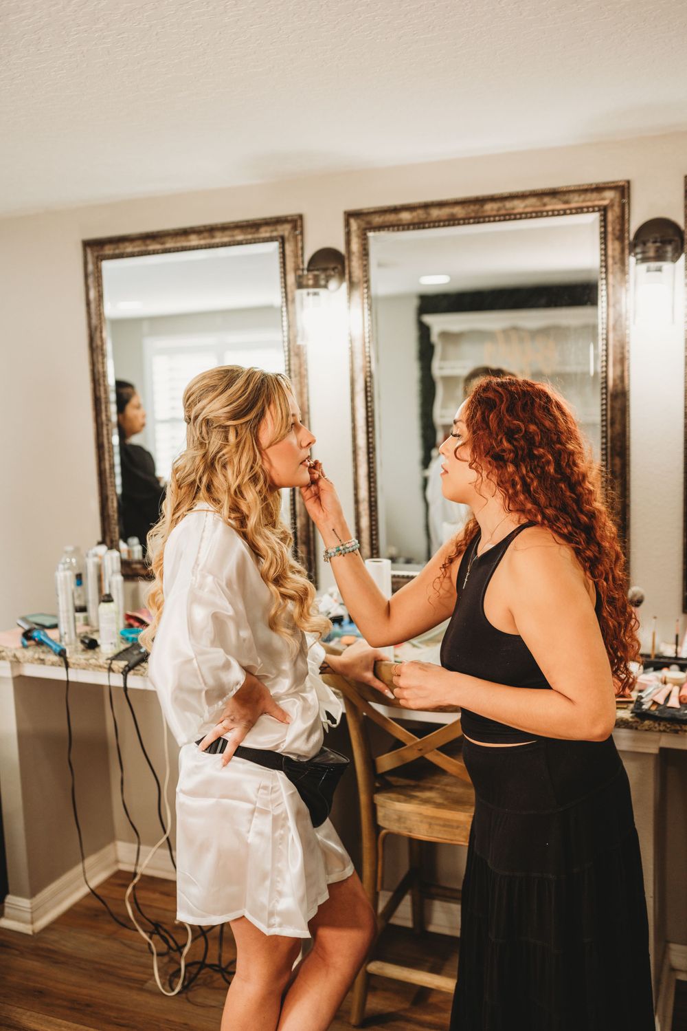 Getting ready for a wedding as stylists work on hair and makeup in a dressing room with illuminated mirrors.